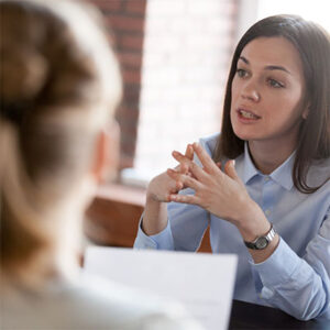 Woman in business meeting, elbows on table, fingers tented slightly as she leans in to earnestly make a point to others sitting across the table from her.