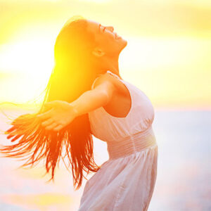 Close-up of a woman standing on a beach with her arms flung happily back as she leans forward with a look of bliss on her face and a sunrise behind her.