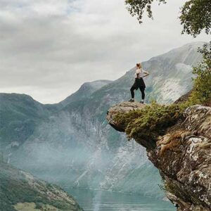 Woman standing atop a cliff, hands on hips, after climbing up. She is looking at a dramatic view of a mountain range.