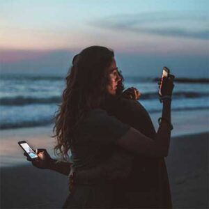 Two people embracing while staring at smart phones over each other's shoulders.